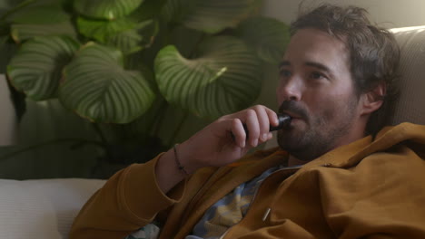 relaxed caucasian young man smoking an electronic cigarette device on couch, close-up shot