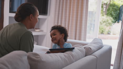 Madre-Con-Hijo-Jugando-O-Transmitiendo-A-Una-Tableta-Digital-Sentados-Juntos-En-Un-Sofá-En-Casa