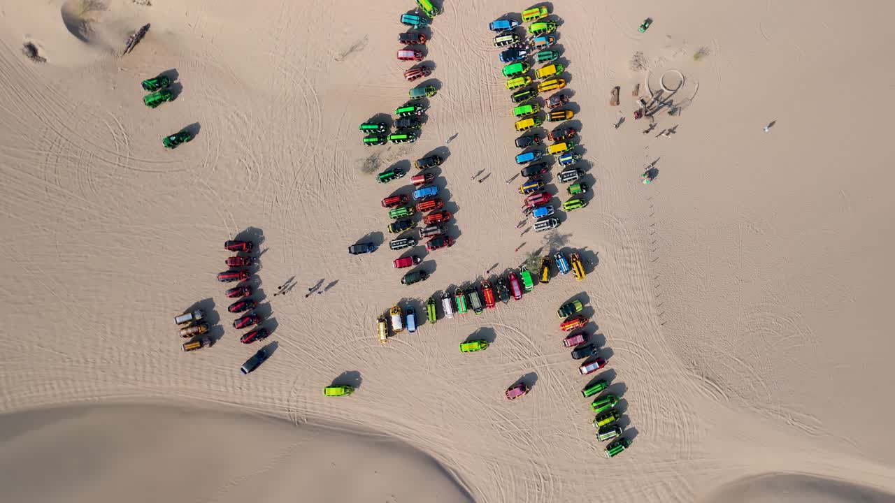 Aerial rising top down shot of Huacachina sand dunes with vibrant dune buggies arranged in patterns for tourists riding in desert Peru