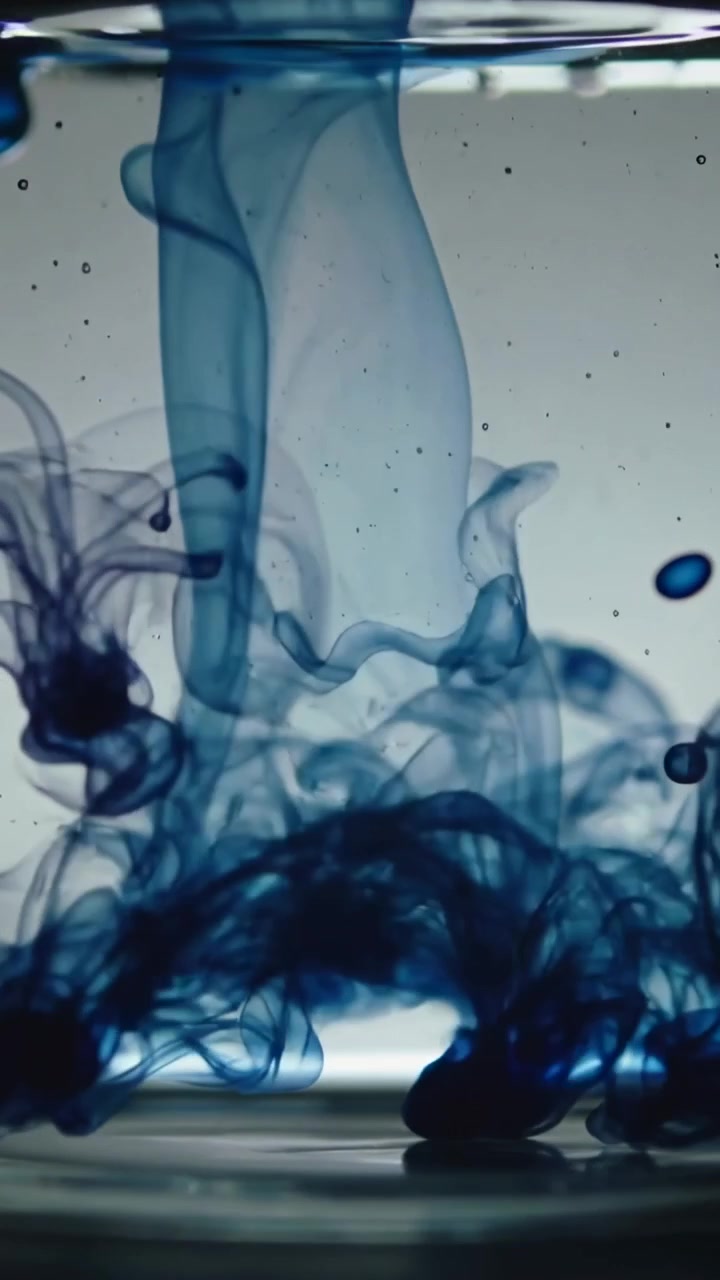 Vertical video: Dropping blue dye forming curling plumes in glass vessel with water to show mixing