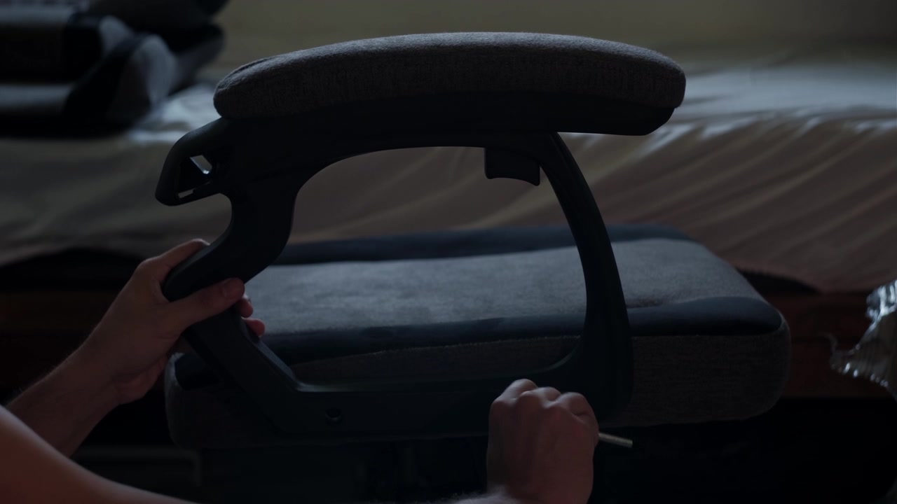 Medium shot of hands installing a gaming chair armrest indoors during furniture assembly with dim lighting and visible tool use