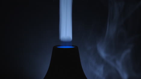 a dark room with a turned on ultrasonic diffuser releasing white smoky aromatic air from essential oil - close up shot