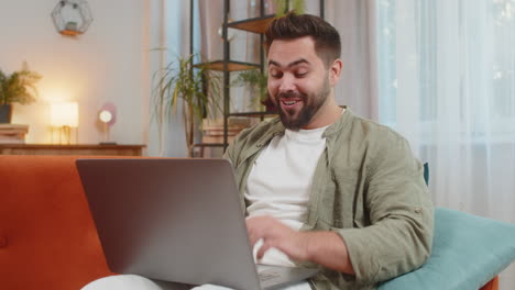 surprised caucasian brunette man using laptop computer sitting on sofa couch at home apartment