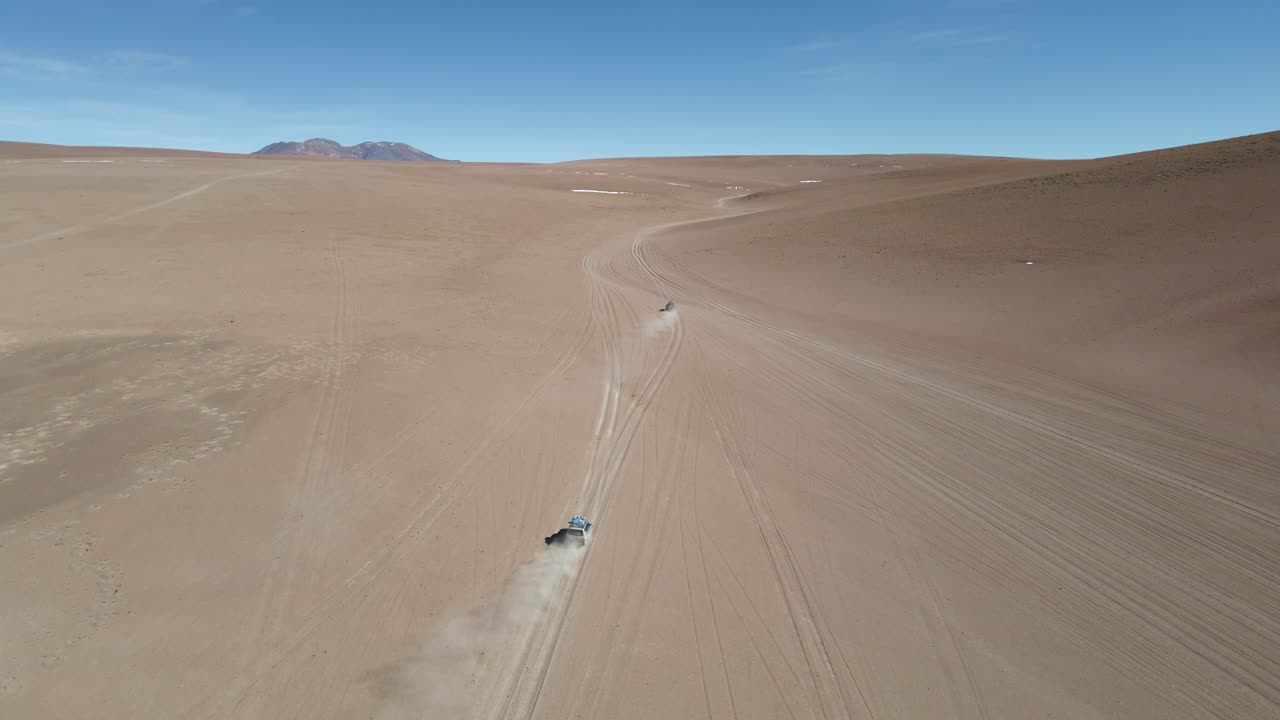 Sweeping drone over wide desert plain in Bolivia shows ridgelines and arid landscape, tracking follows cars with dust rising behind