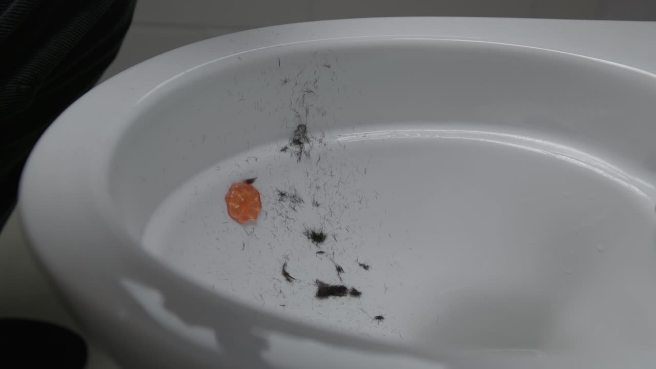 Close-up of hair clippings in a white toilet after shaving a head. A razor is tapped out, loose hairs falling against clean porcelain in a simple bathroom setting. Caucasian male shaving head