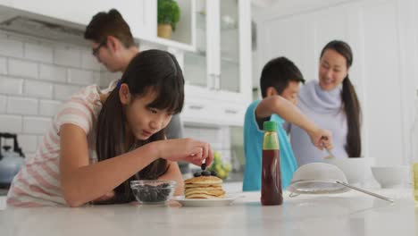 Happy-asian-parents-in-kitchen-cooking-with-son-and-daughter,-daughter-putting-berries-on-pancakes