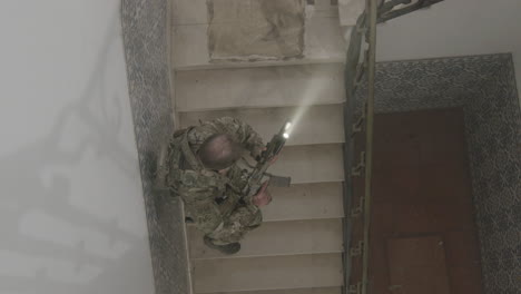 long shot of a focused soldier with scratches on face climbing stairs slowly with rifle and flashlight