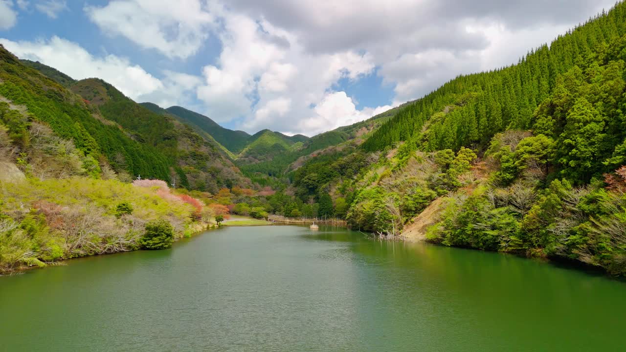 A stunning aerial drone shot slowly flies over a calm, green lake nestled in a vast mountain valley. The forested hillsides show vibrant spring colors under a partly cloudy sky.