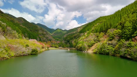a stunning aerial drone shot slowly flies over a calm, green lake nestled in a vast mountain valley. the forested hillsides show vibrant spring colors under a partly cloudy sky.