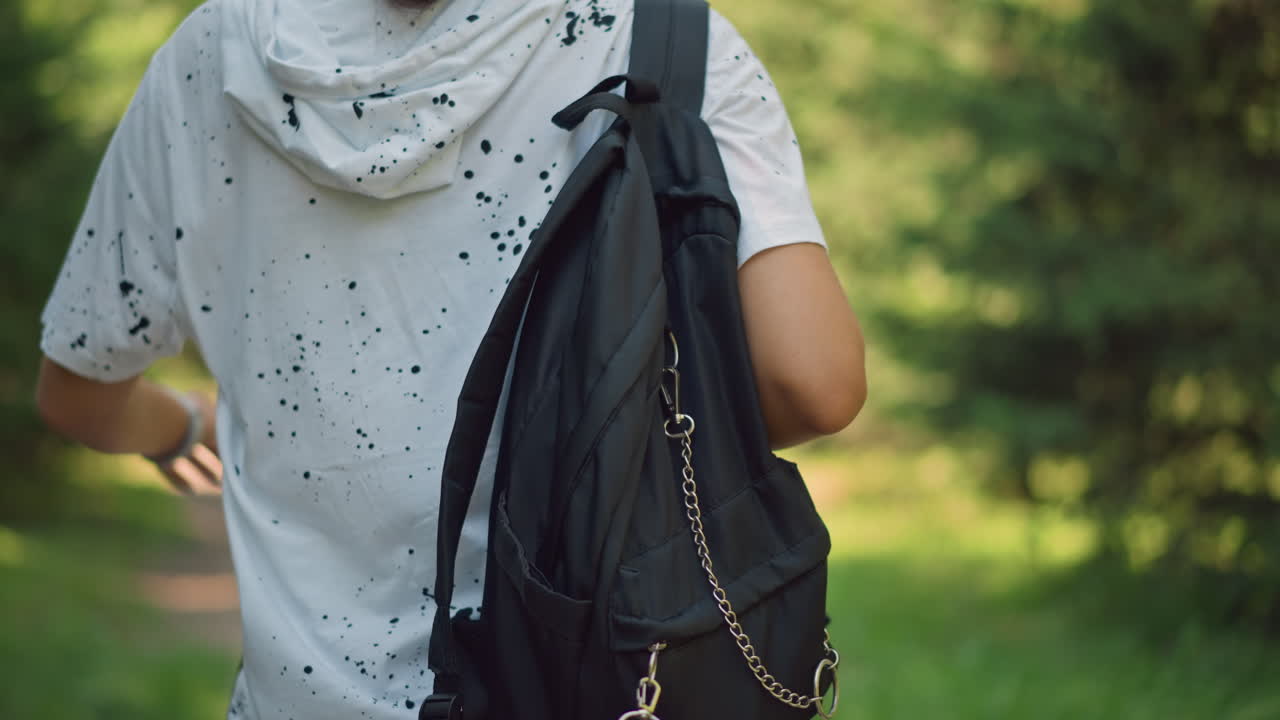 persona joven caminando con mochila al aire libre, primer plano de la espalda de un joven informal que lleva un bolso negro al hombro por un sendero arbolado iluminado por el sol, detalles de una camisa blanca moteada, ritmo veraniego relajado, poca profundidad de campo