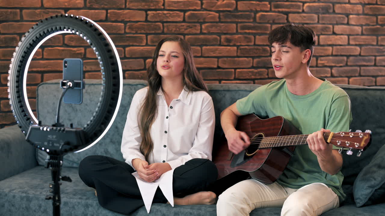 Young man and woman playing on a guitar and singing, shooting themselves on smartphone on a tripod with ring light in a studio