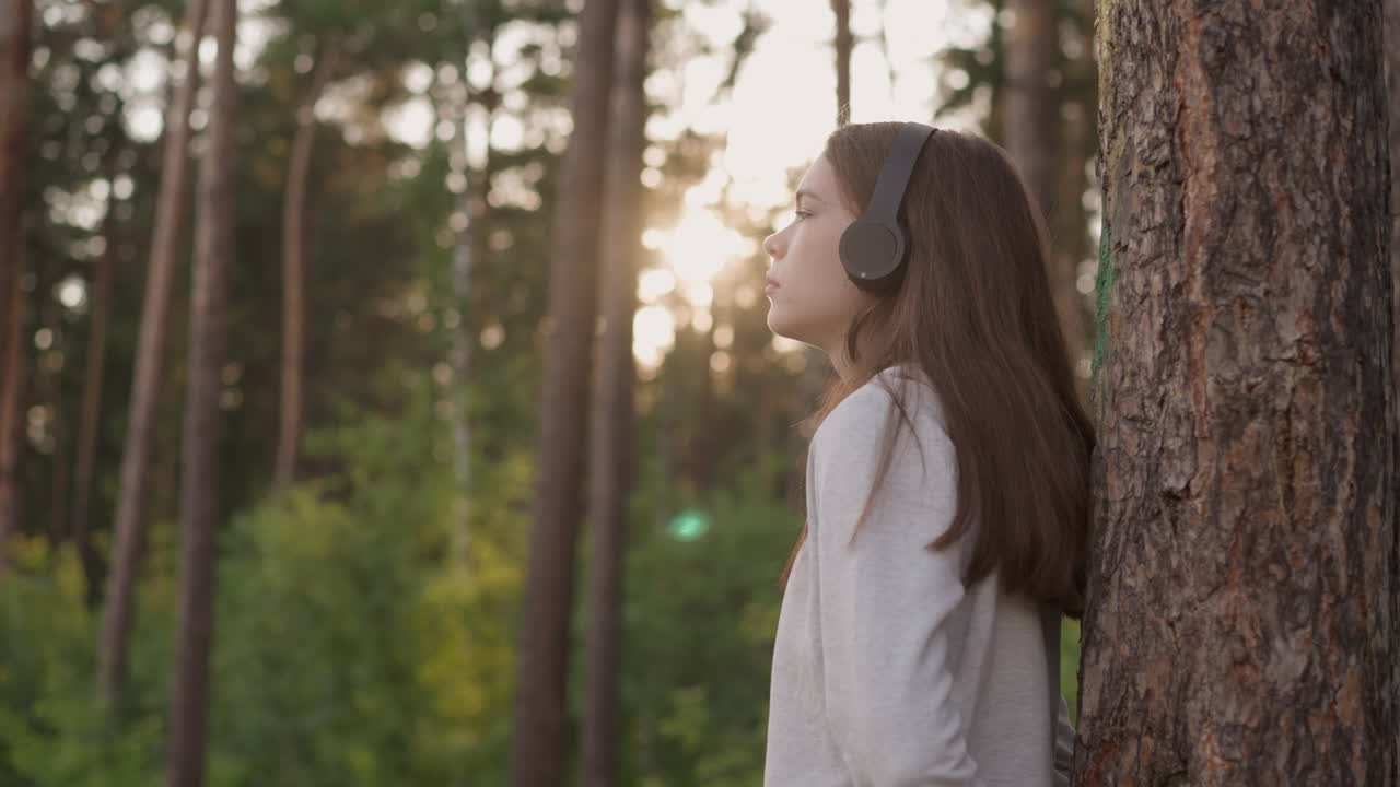 mujer con auriculares se apoya en el tronco de un árbol en el bosque. joven escucha música melancólica pensando en la experiencia amarga al atardecer. sensación de soledad