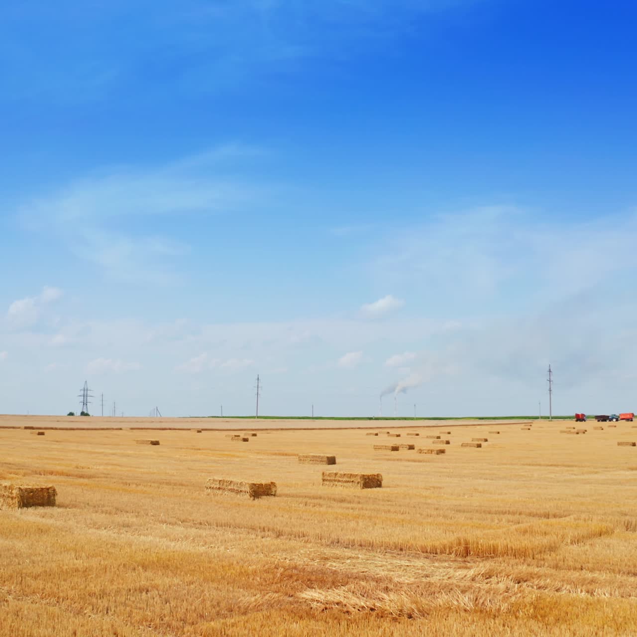 Landbrugsplantage i sommerhøstsæsonen. Rektangulære høballer på marken og landbrugsmaskiner. Blå himmel i baggrunden