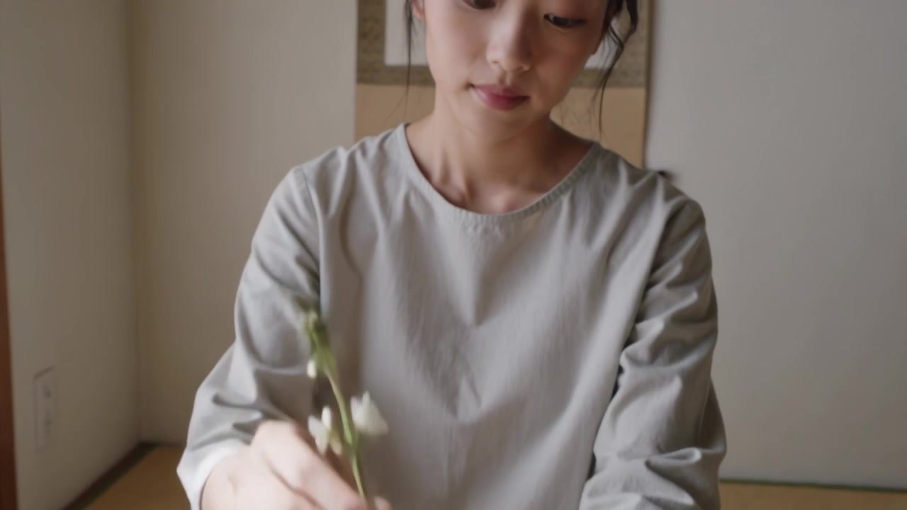 Raccogliendo steli a sinistra, una donna con un top grigio sistema steli bianchi in un cesto in una stanza tatami per l'ikebana.