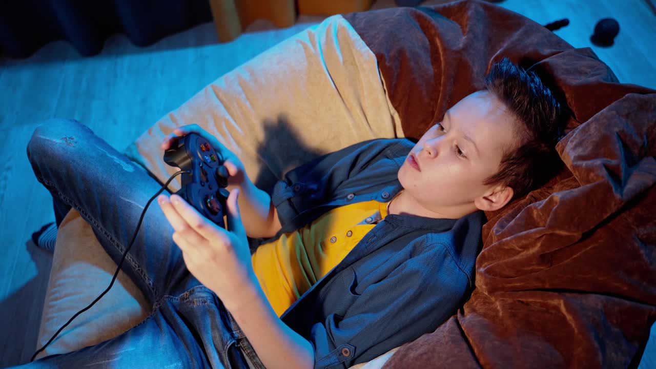 Young gamer playing video games at home. Boy laying in comfortable armchair and managing joystick while playing modern games.