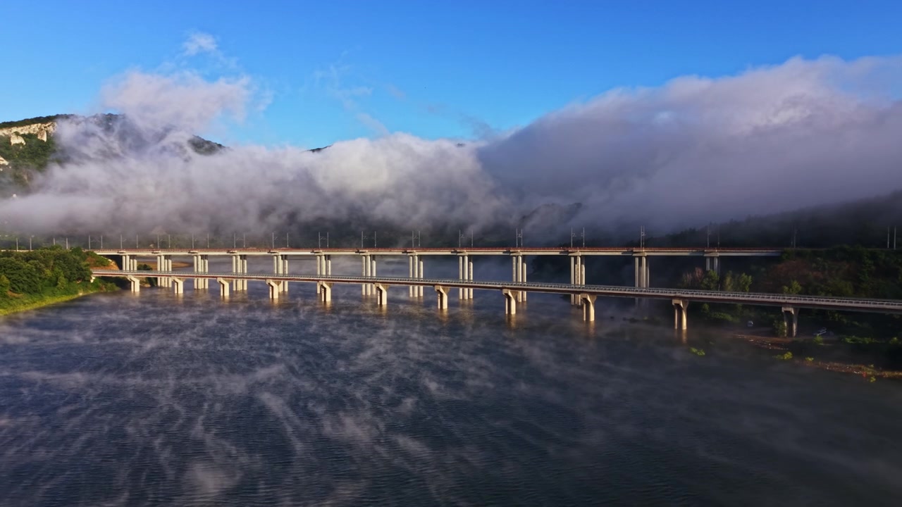 晴れた日の青空の下、川には霧と雲がたなびき、橋が両岸をつないでいる。
