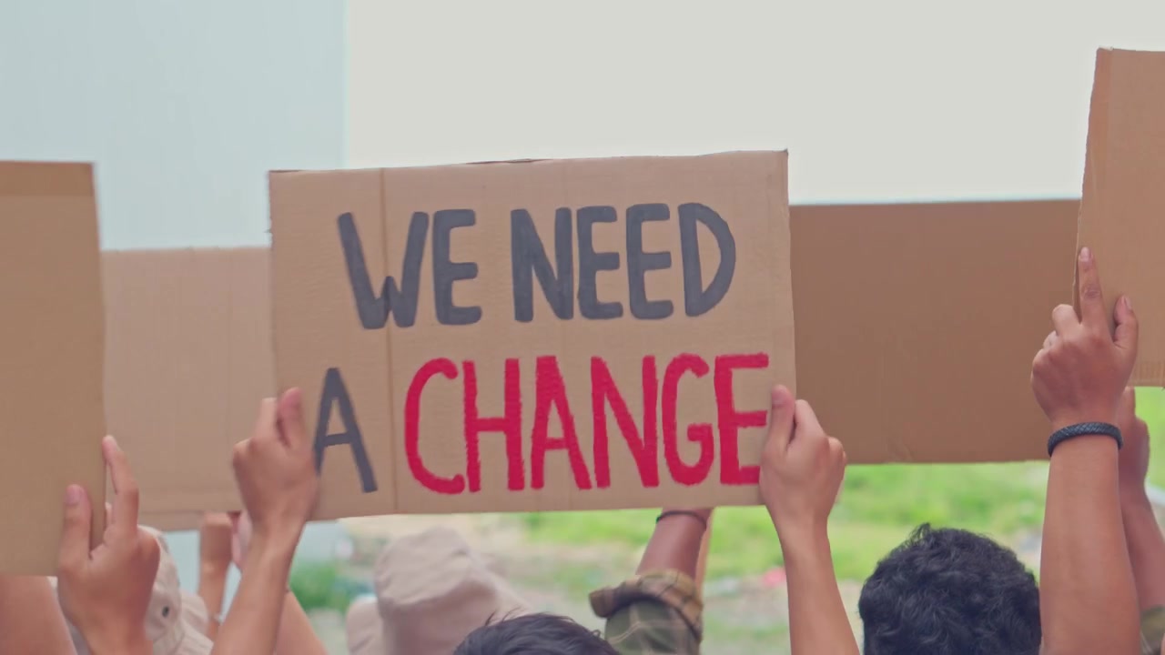 Crowd of protesters holding signs high, featuring "We Need A Change" during demonstration