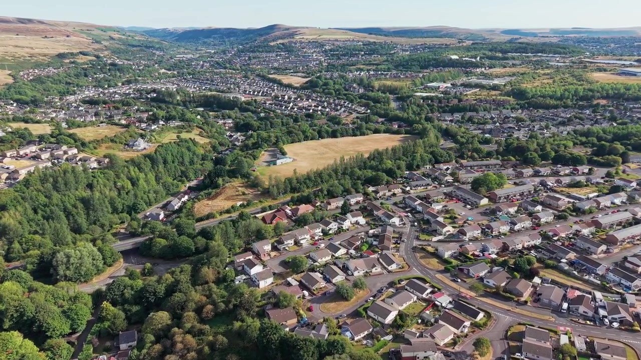Vista aérea da cidade de Ebbw Vale no Sul do País de Gales, com conjuntos habitacionais residenciais espalhados pelo fundo do vale e pelas colinas verdes circundantes durante o tempo seco de verão
