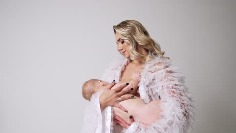 beautiful woman with long blond hair wearing white robe waving a baby in hands. mother and child concept. white backdrop.