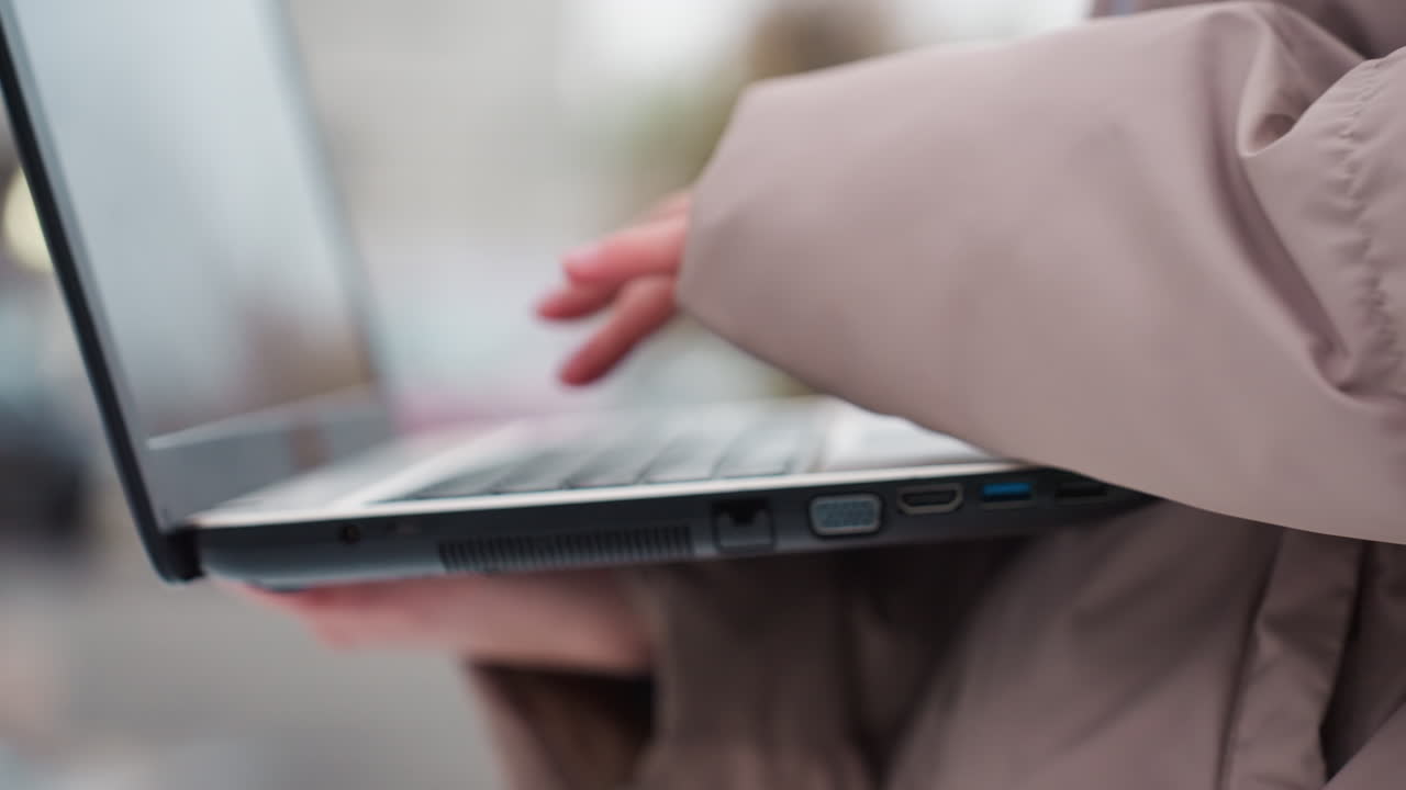 Close-up view of light skin woman using laptop while wearing winter jacket outdoors, hands actively typing with blurred background, showing concentration, and use of technology in cold environment