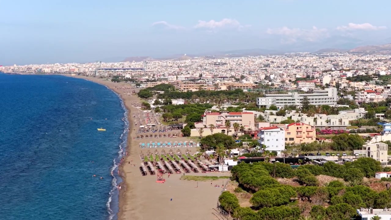 Vista aérea de drone da longa praia de areia com guarda-sóis e espreguiçadeiras que se estende em direção à grande cidade costeira, Ammoudara, Grécia