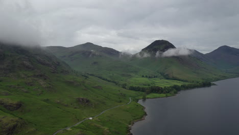 Aerial-view-of-green-mountains-at-the-side-of-a-lake,-road-running-along-lake-side,-cloudy-day