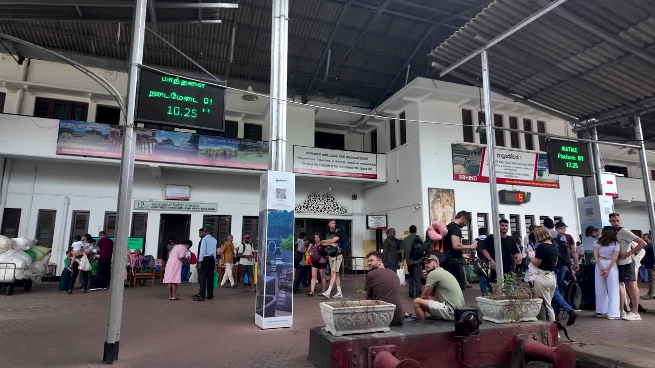 Kandy train station with travelers and digital information boards displaying schedules. The platform is crowded with people waiting for their trains in Sri Lanka.