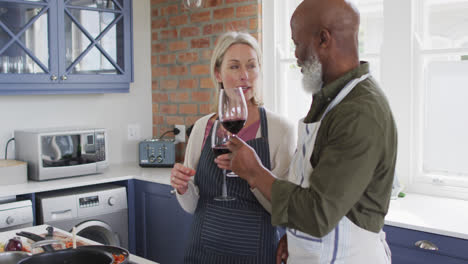una pareja de adultos mayores de raza mixta con delantales brindando copas de vino mientras cocinan en la cocina de casa