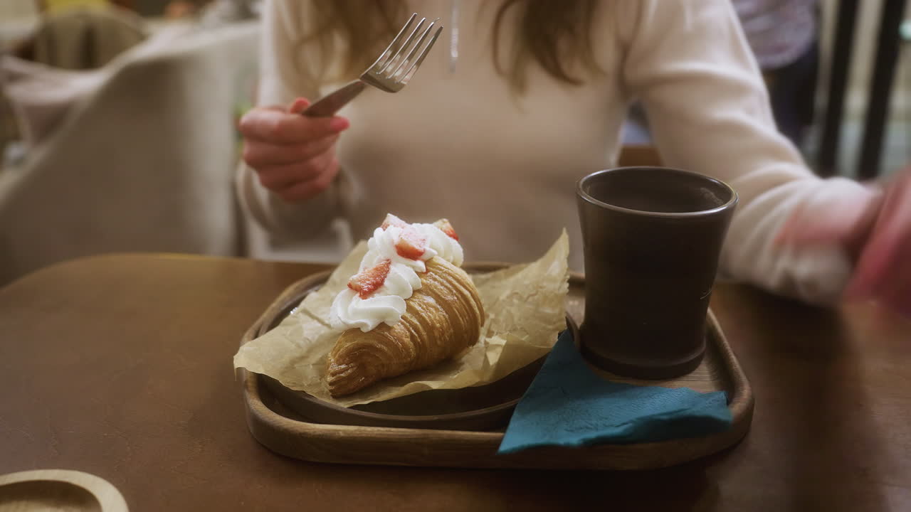 Croissant with whipped cream and strawberries, paired with a cup of coffee, placed on table in cozy cafe. Female in beige shirt, face not visible, enjoying breakfast in a relaxed atmosphere