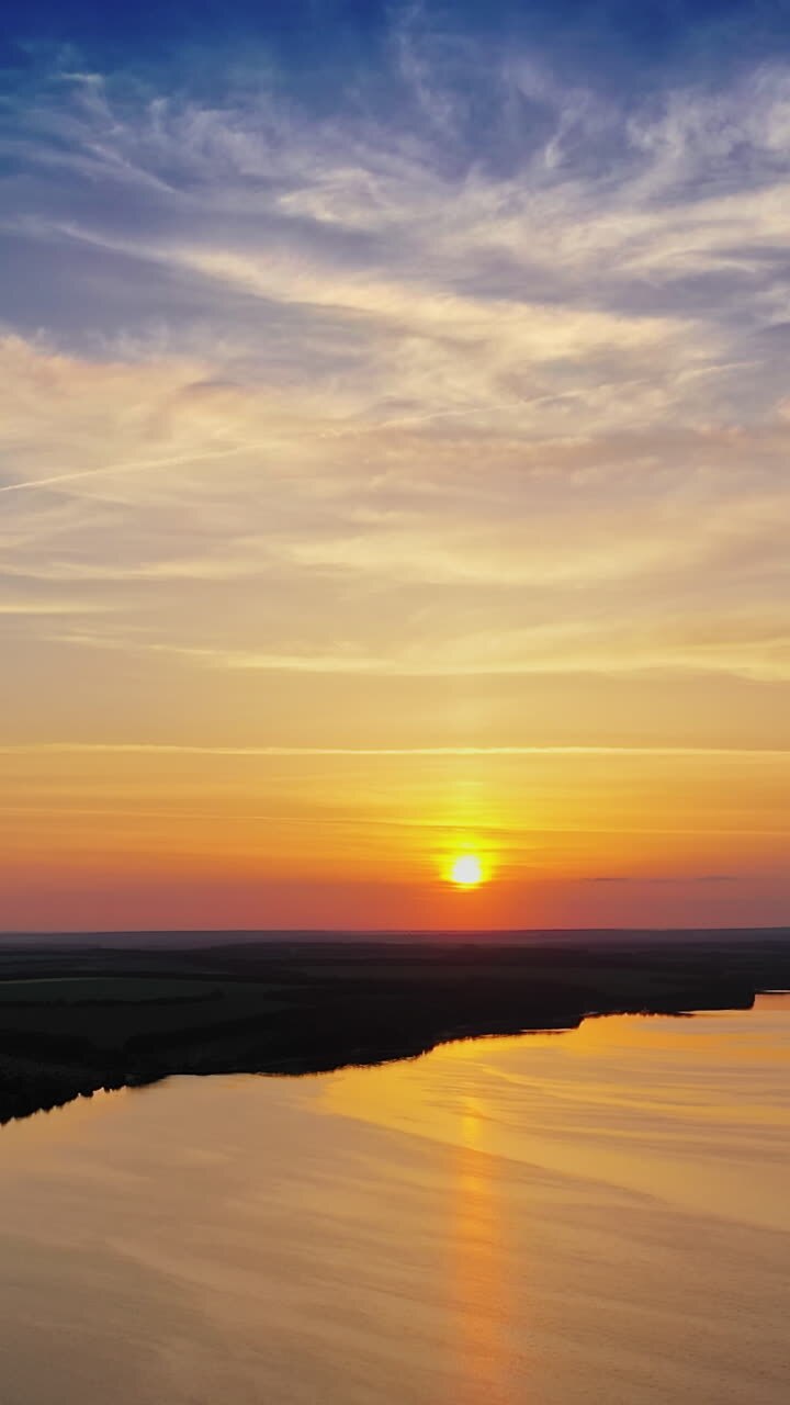 Amazing orange sunset over the river. Magic aerial view of evening sun among clouds. Golden path of setting sun on the calm river in nature. Slow motion camera. Vertical video