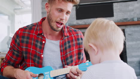 dad sits playing ukulele and singing with toddler, close up