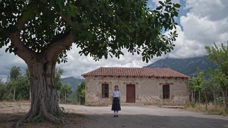schoolgirl in uniform is standing in the schoolyard of a rural school building under the shade of a large tree, with mountains in the background, on a cloudy summer day