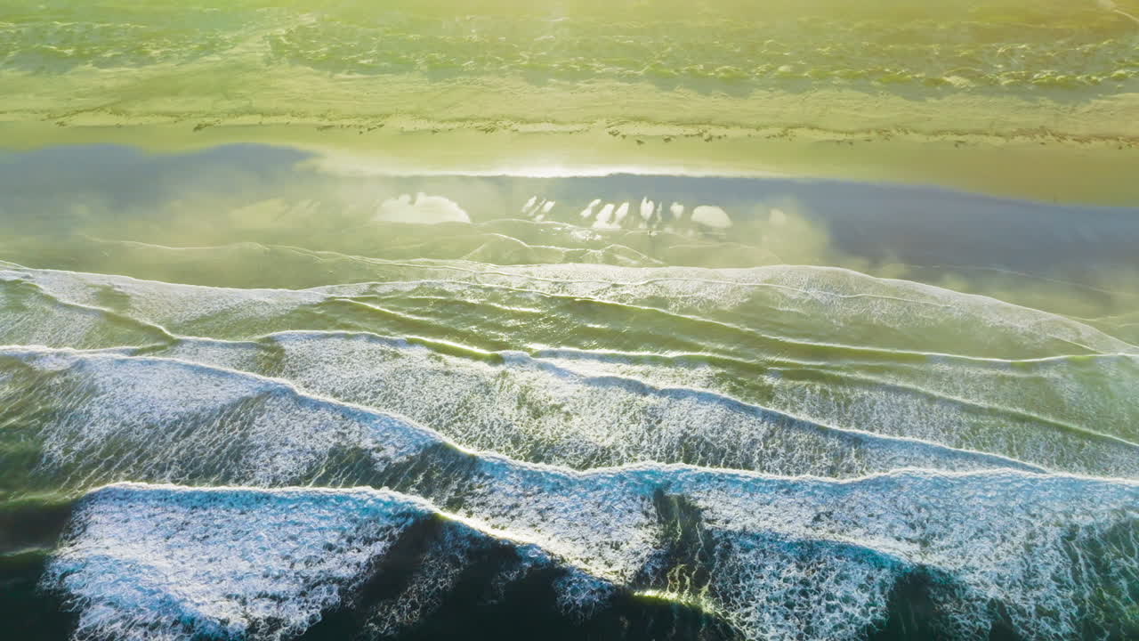 White foamy little waves splashing on the sandy beach at daytime. Bright sun lighting the coast of the ocean. Top view.