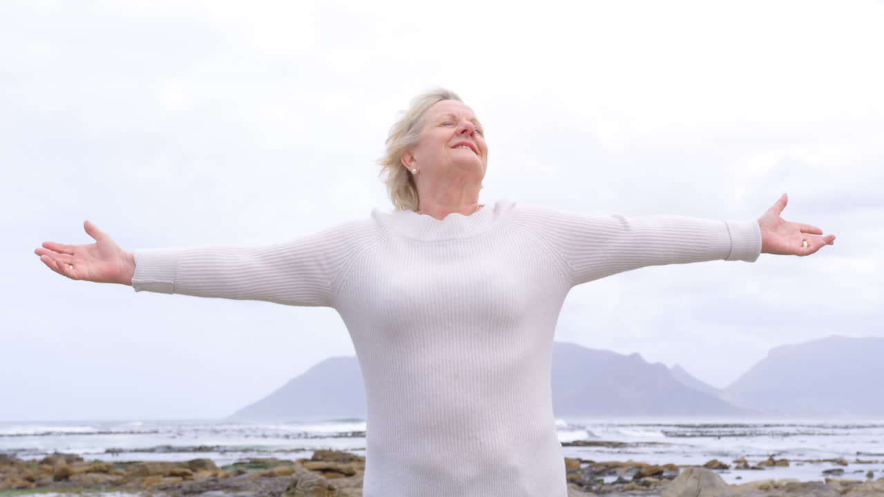4k 해변의 바위 위에 팔을 펴고 서 있는  ⁇ 은 백인 노인 여성의 앞면