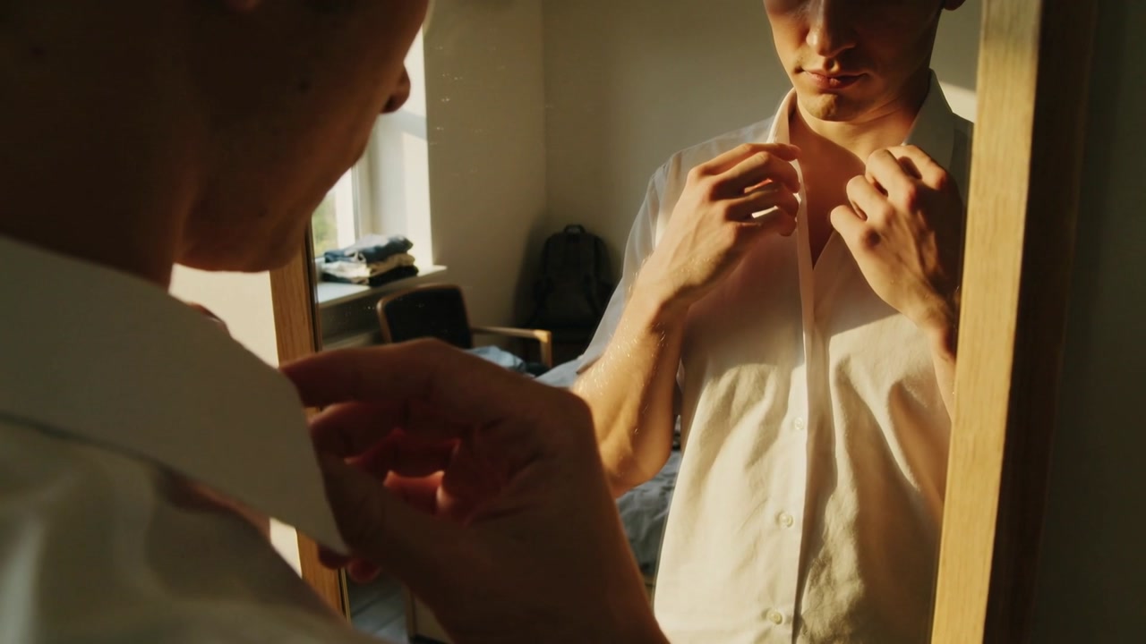 Male student standing in small bedroom, checking mirror and adjusting white shirt collar for work