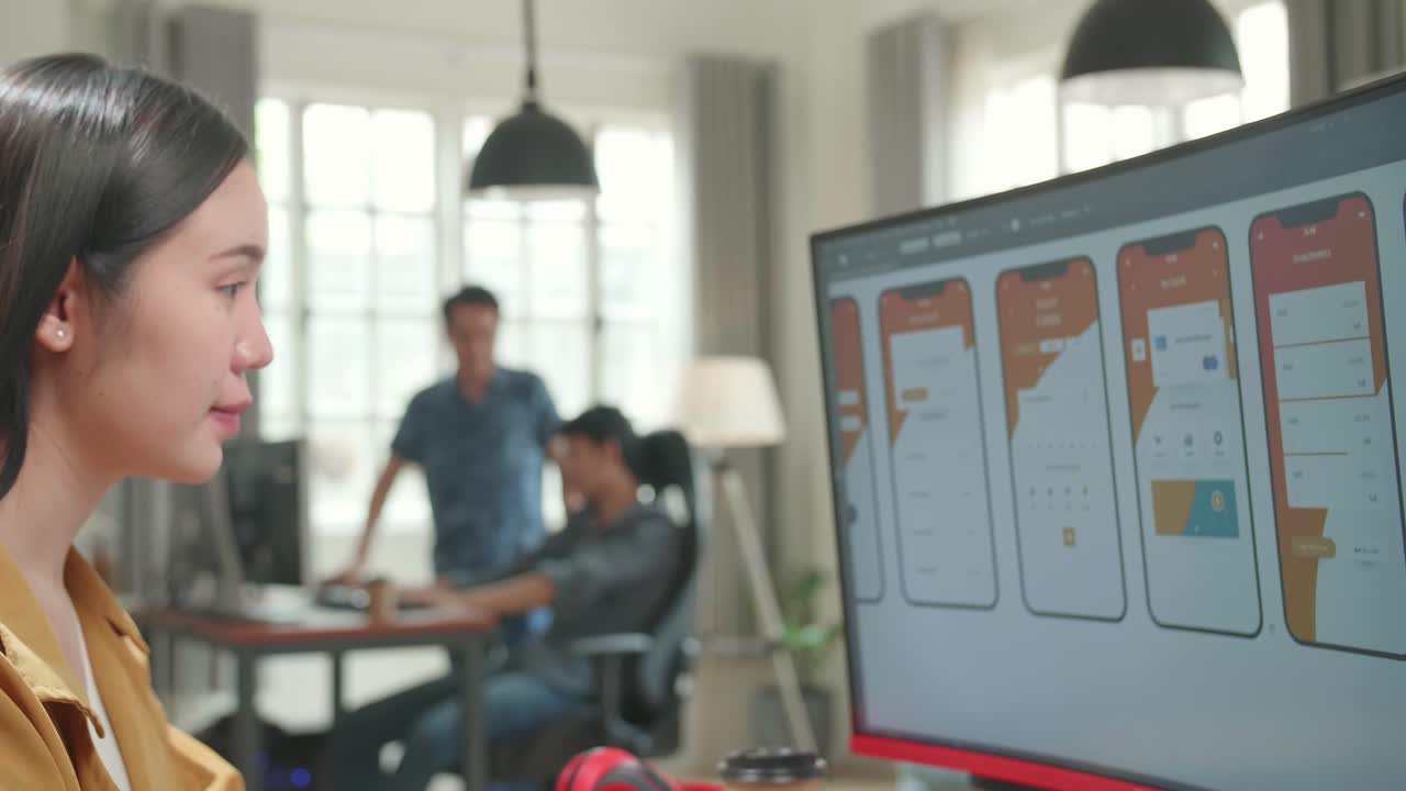 Asian Female Mobile Application Developer Works With Graphics On Her Personal Computer In Office. She Turns And Warmly Smiles Into The Camera