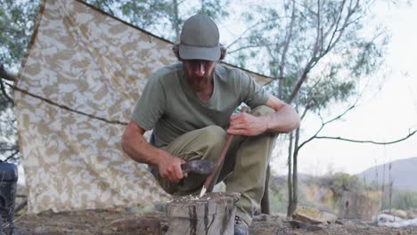 Bearded-caucasian-male-survivalist-sharpening-stick-with-machete-at-camp-in-wilderness