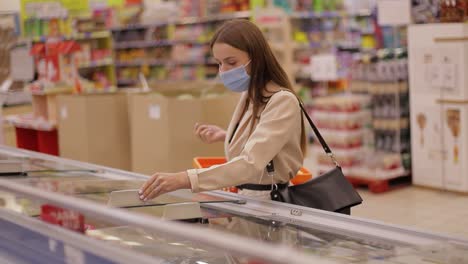 a young woman in a medical mask buys food in a supermarket during the covid-19 coronavirus epidemic. the woman stores food during quarantine and self-isolation. social video