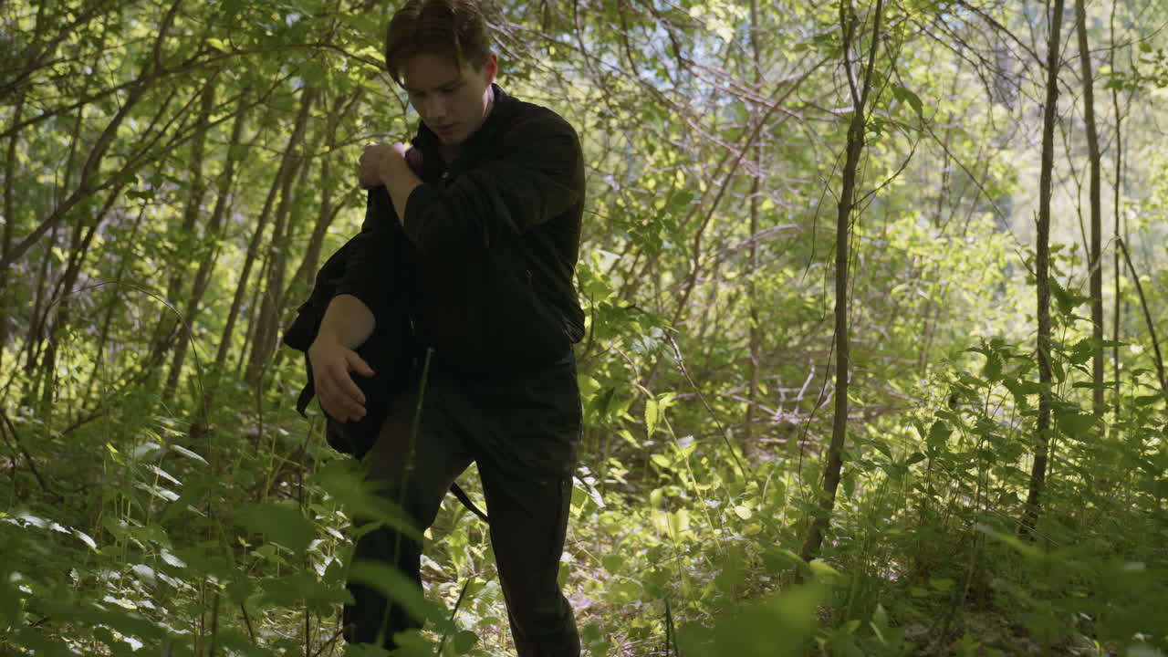 Tourist dressed in black outdoor outfit cautiously navigates dense forest, clearing branches with hand while kneeling to access backpack, surrounded by bright green foliage and filtered sunlight