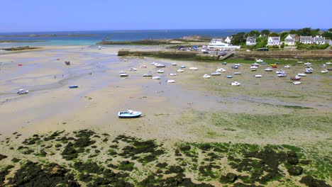 drone view of a coastal area in brittany, france, showing boats moored on sandy, rocky beach at low tide. exposed seabed reveals seaweed patches with houses and blue sea in the background.