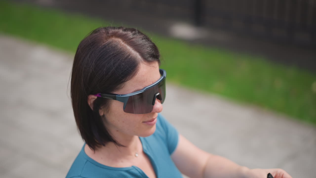 Woman fastening helmet strap before ride, checking visor and handlebar grip on driveway, teal top, short bob hair, reflective glasses, closeup of clasp and buckle, focused expression, practical calm