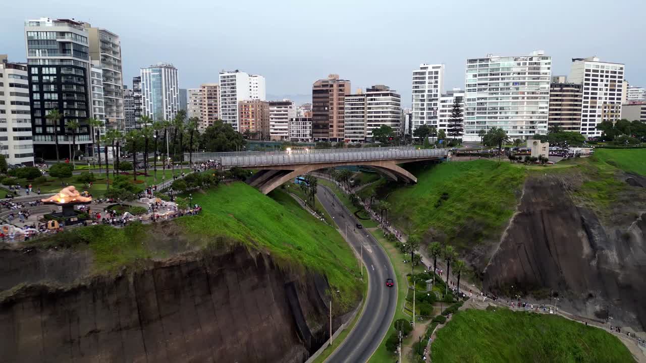 Rekaman dolly mundur udara menunjukkan jembatan kota beton di Amerika Selatan yang menghubungkan dua jurang berumput pesisir, dikelilingi oleh bangunan, taman dengan pohon palem, dan orang-orang yang berjalan.