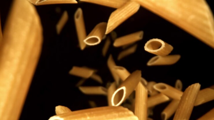 Pezzi di pasta penne cruda che cadono al rallentatore su sfondo nero.