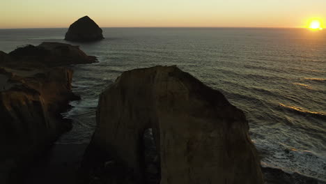 Roca-De-Pajar-En-La-Distancia-Pasando-Por-El-Arco-Erosionado-De-Arenisca-Del-Cabo-Kiwanda-Al-Atardecer,-Aéreo