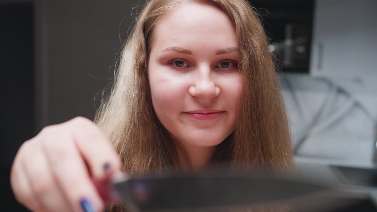 Zoom in portrait elegant woman in modern kitchen smiling, playfully turning knife toward camera, blowing kisses with polished nails visible, soft light on face, casual indoor background