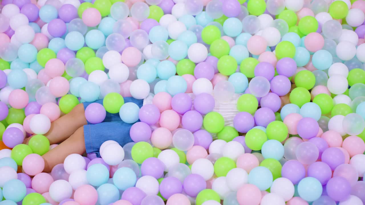 Happy little girl lying in a ball pit as colorful plastic balls fall on top of her in a fun kids playroom - top down slow motion