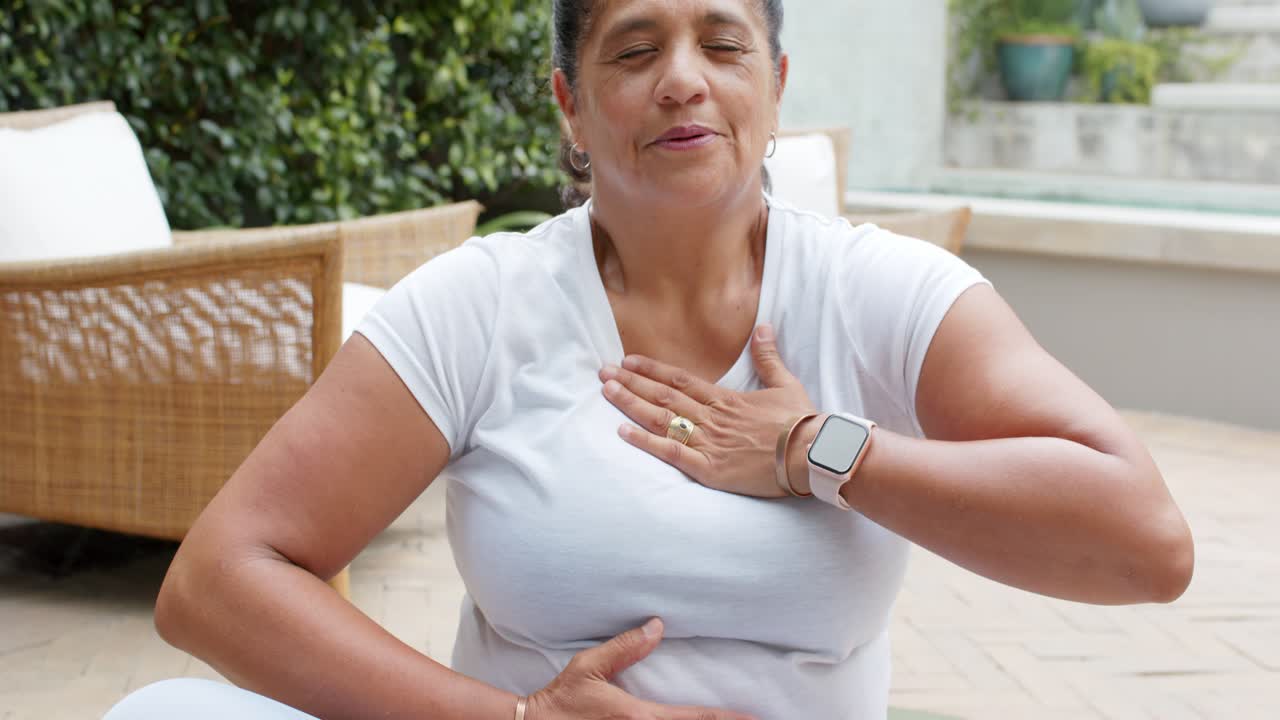 Câmera se afastando, mulher afro-americana madura em colchonete respirando com as mãos no peito e abdômen para se acalmar