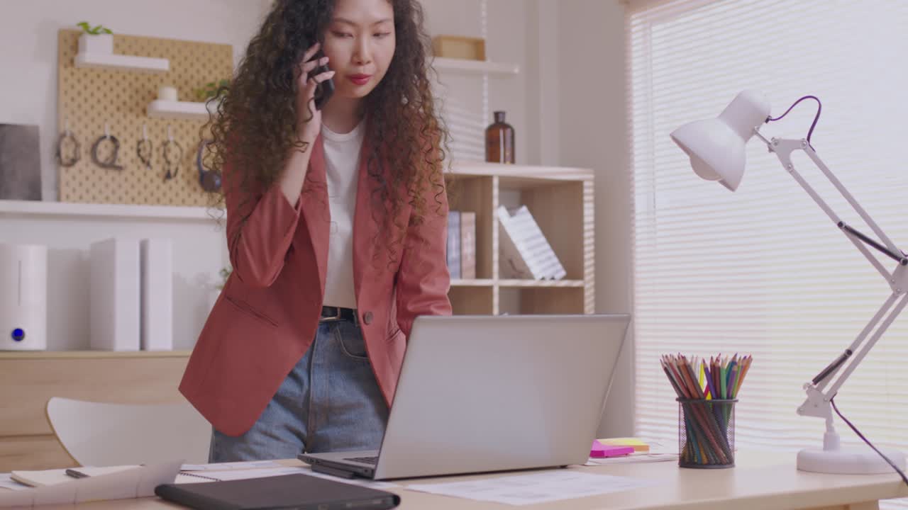 la mujer diseñadora asiática usa su teléfono inteligente para comunicarse, trabajar en proyectos y papeles, y trabajar desde casa en un escritorio con una computadora portátil.