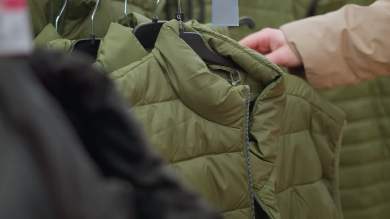 A close-up of a hand in a brown jacket reaching for a green jacket from a clothing rack in a mall. highlights the texture of the puffy jacket and the casual nature of shopping
