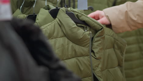 un primer plano de una mano con una chaqueta marrón alcanzando una chaqueta verde de un estante de ropa en un centro comercial. destaca la textura de la chaqueta hinchada y la naturaleza casual de las compras